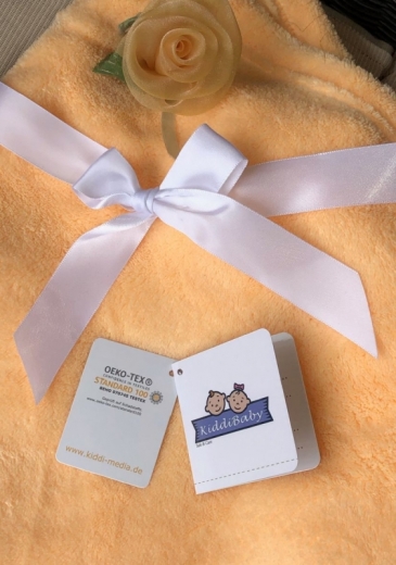 Morbida copertina per neonati arancione con fiocco in raso bianco e delicata rosa in tessuto giallo chiaro. Sulla copertina sono appesi due cartellini: il certificato Oeko-Tex Standard 100 e l'etichetta KiddiBaby. Sono garanzia di sicurezza e comfort.