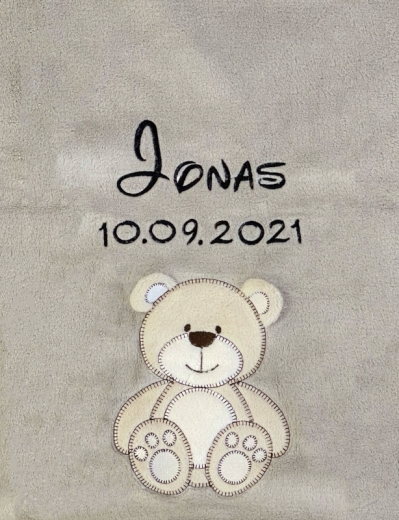 Coperta per neonati grigia, morbidissima e ricamata: in alto al centro il nome Jonas in corsivo nero, sotto la data di nascita 10/09/2021. Sotto, un orsacchiotto color beige con contorni marrone scuro e un'espressione sorridente.