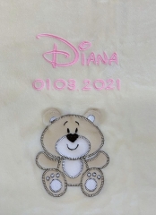 Primo piano di una copertina per neonati beige con ricamo: nome «Diana» in corsivo rosa e data di nascita «01.03.2021». In basso, applicazione a forma di orsacchiotto beige sorridente con zampe bianche.