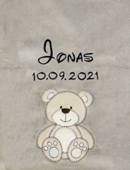 Coperta per neonati grigia, morbidissima e ricamata: in alto al centro il nome Jonas in corsivo nero, sotto la data di nascita 10/09/2021. Sotto, un orsacchiotto color beige con contorni marrone scuro e un'espressione sorridente.